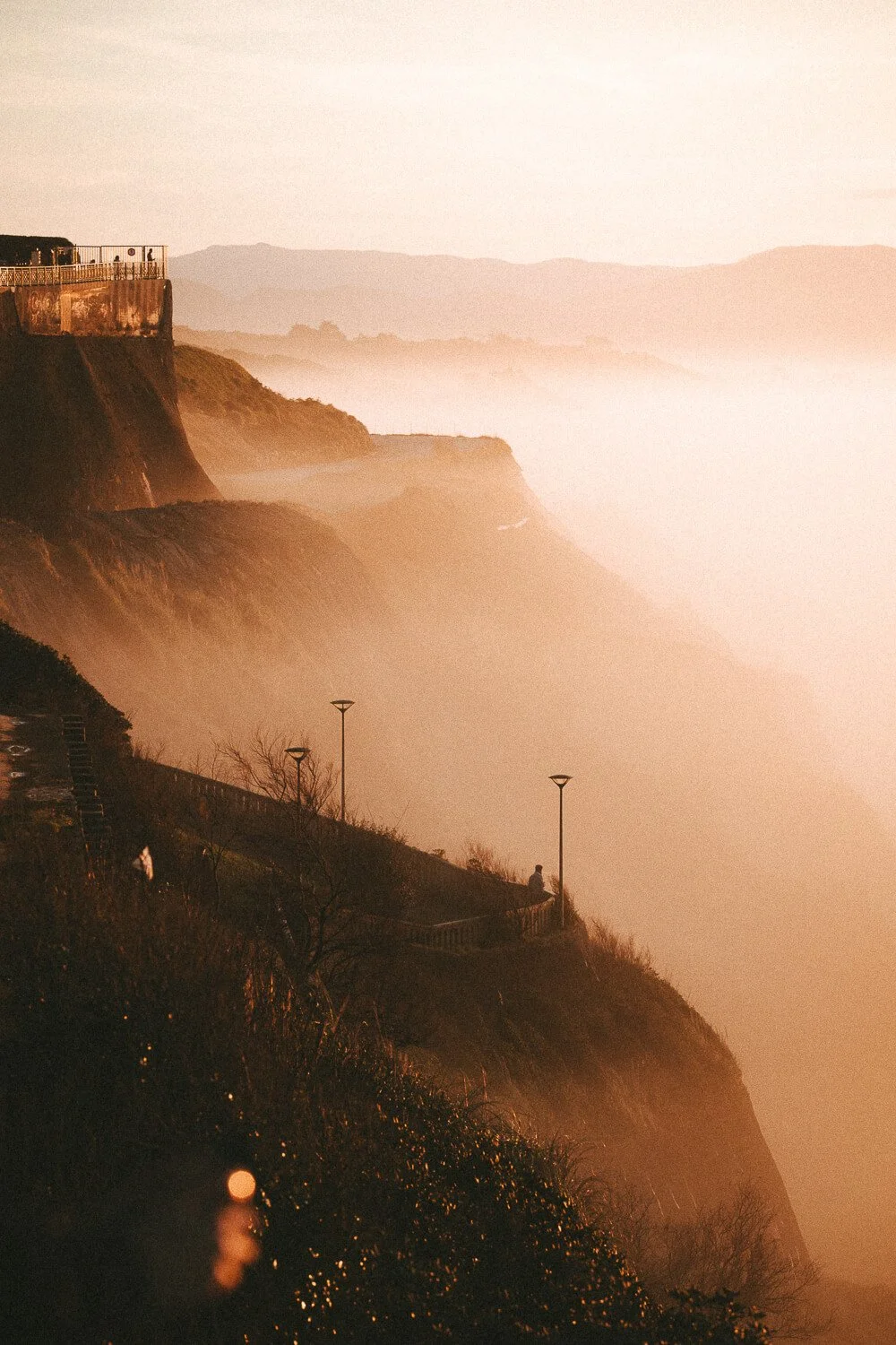Sunset over rugged cliffs along a foggy coastline, with a person sitting on a railing and street lamps on a pathway.