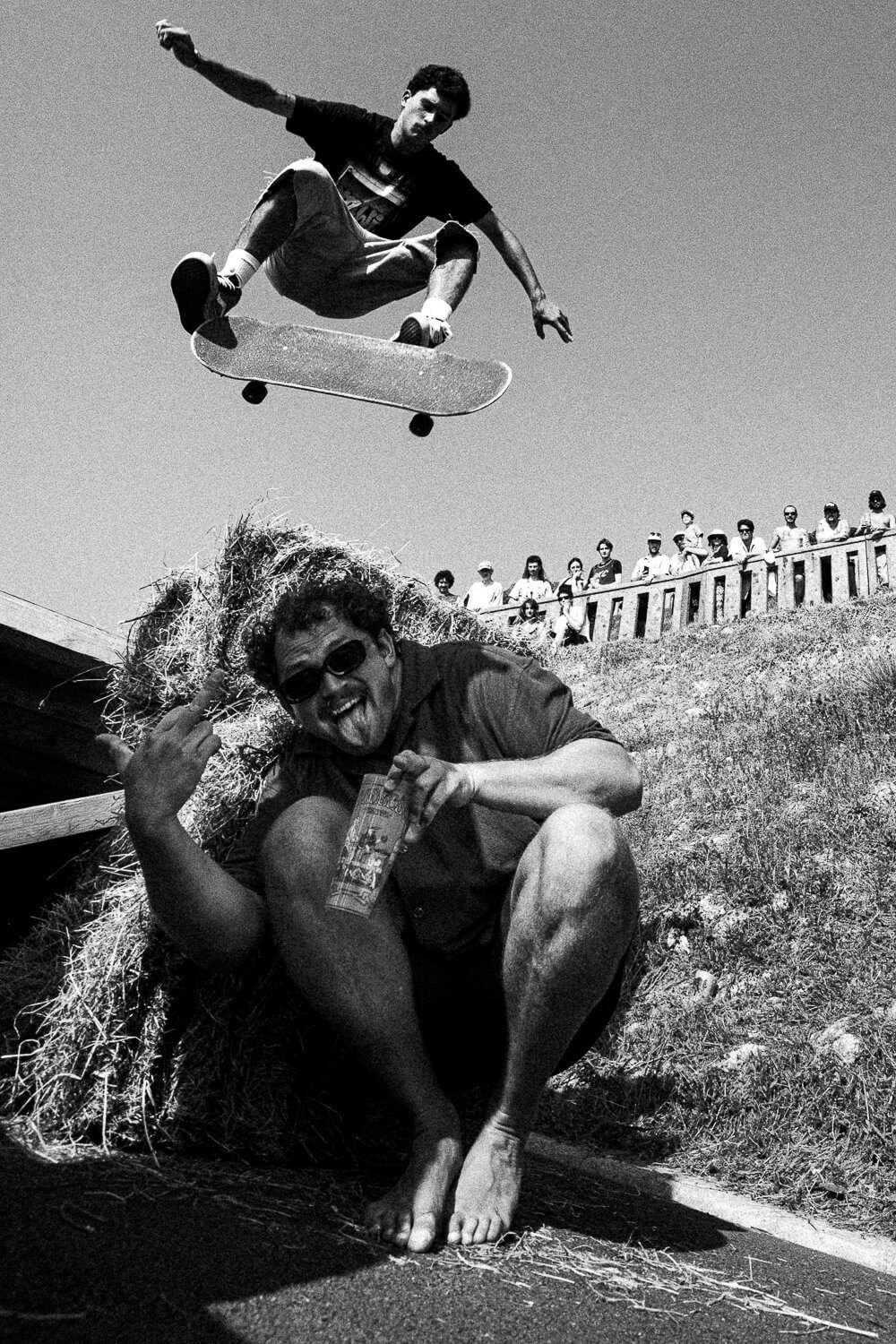 A skateboarder in mid-air performing a trick above a person sitting on hay, who is smiling and holding a drink, with a crowd of spectators watching from above.