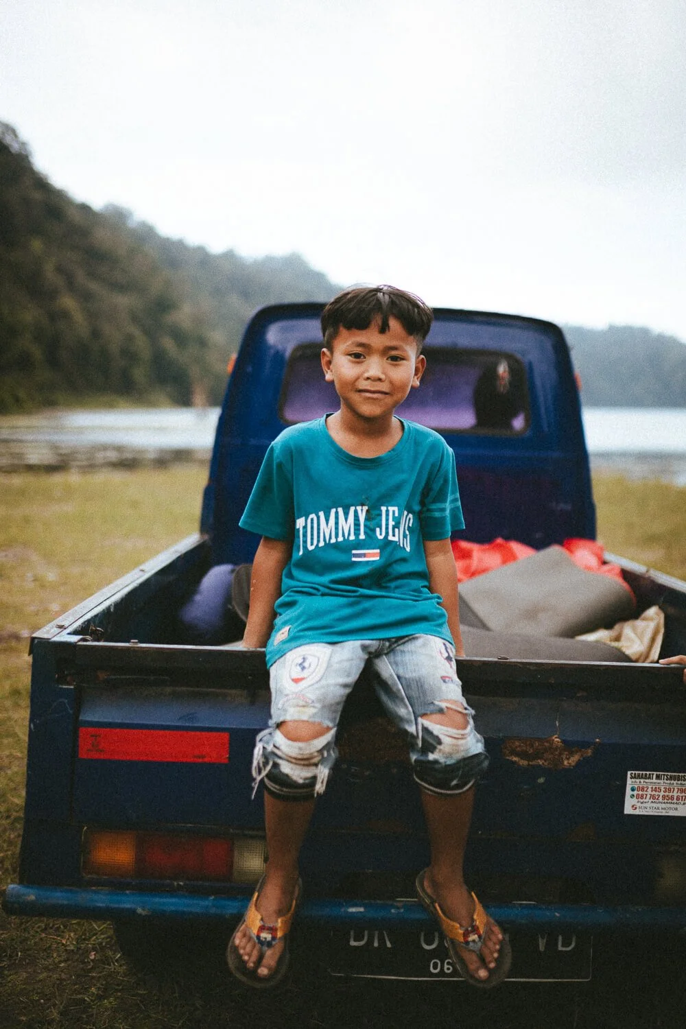 Un garçon assis à l'arrière d'une camionnette bleue, avec un paysage verdoyant et une rivière en arrière-plan.