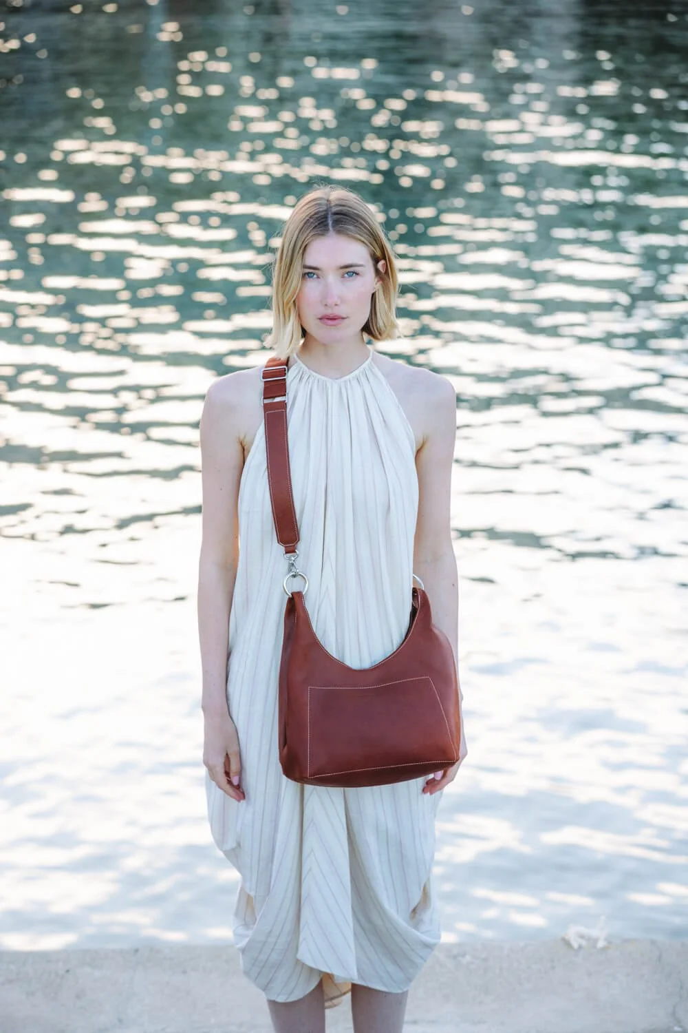 A young woman with blonde hair wearing a white pleated dress and carrying a brown leather shoulder bag stands on a sandy beach near a body of water, with the sunlight reflecting on the water surface in the background.