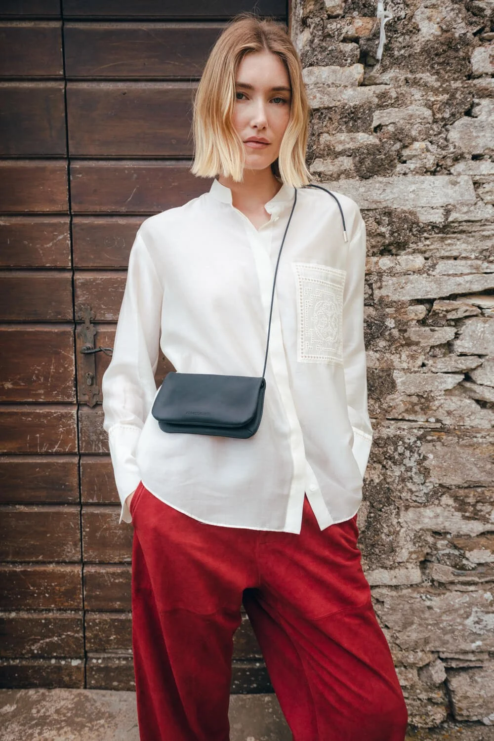 A young woman with blonde hair styled in a shoulder-length bob, wearing a white button-up shirt with embroidery on one pocket, red pants, and a small black crossbody bag. She is standing outdoors against a background of wooden and stone walls.