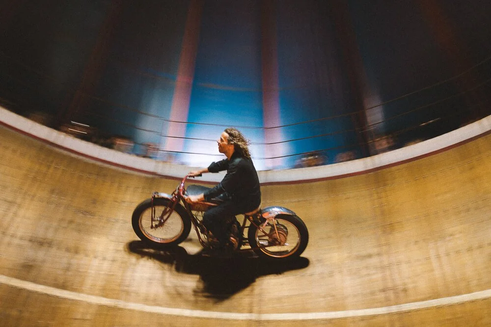 Person riding a bicycle inside a large, circular wooden structure, possibly a velodrome with a curved, enclosed track.