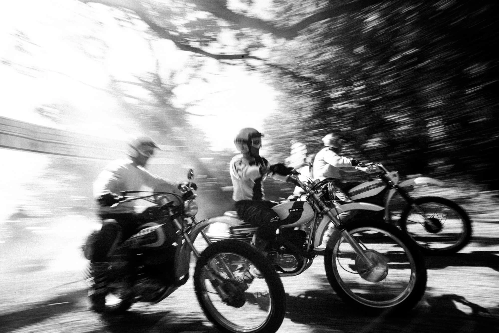 Group of motorcyclists riding at high speed on a road during daytime, with motion blur and sunlight filtering through trees.