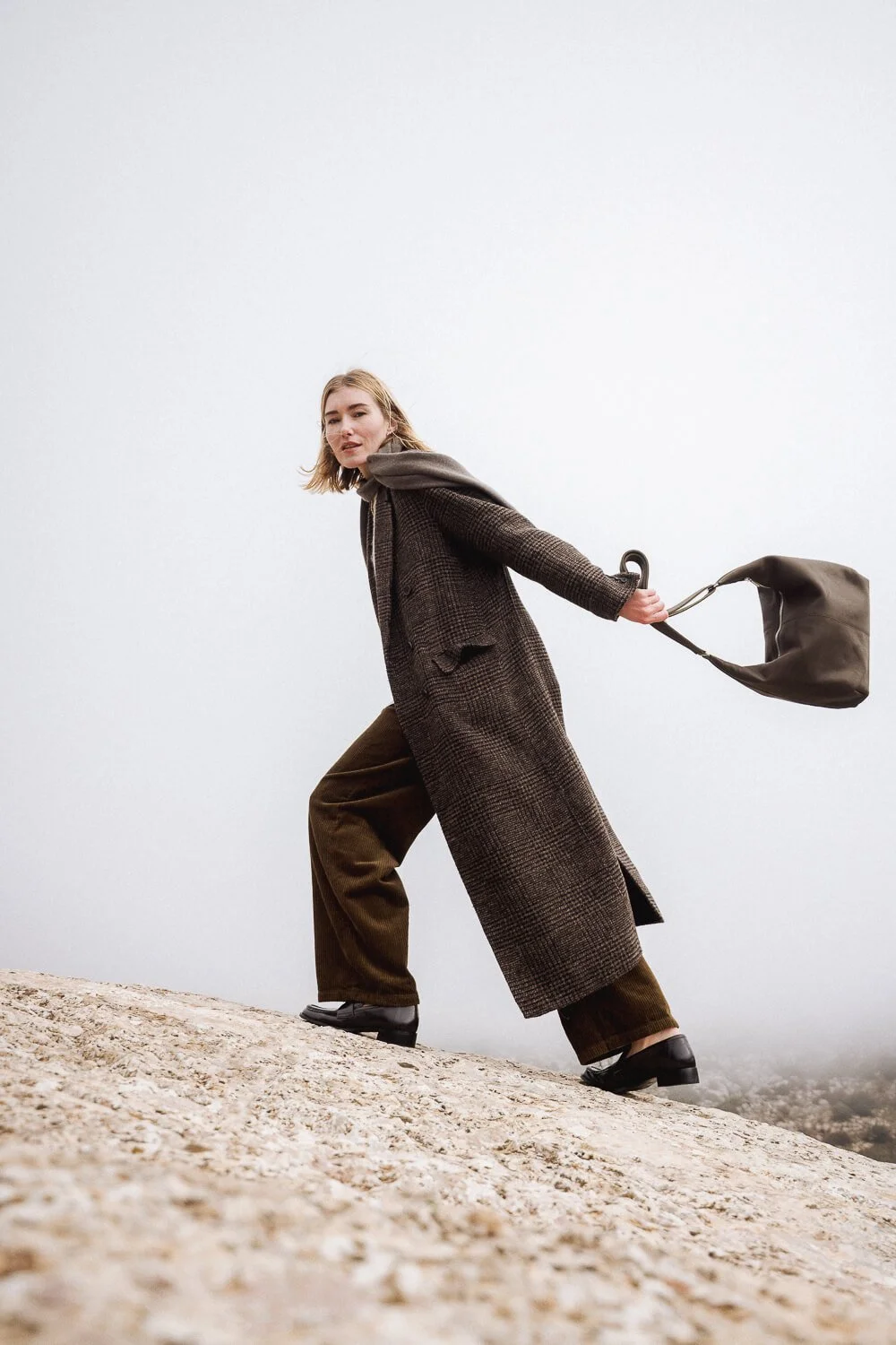 A woman in a long coat and loose pants is leaning forward on a rocky surface, holding a handbag behind her, with a cloudy sky backdrop.