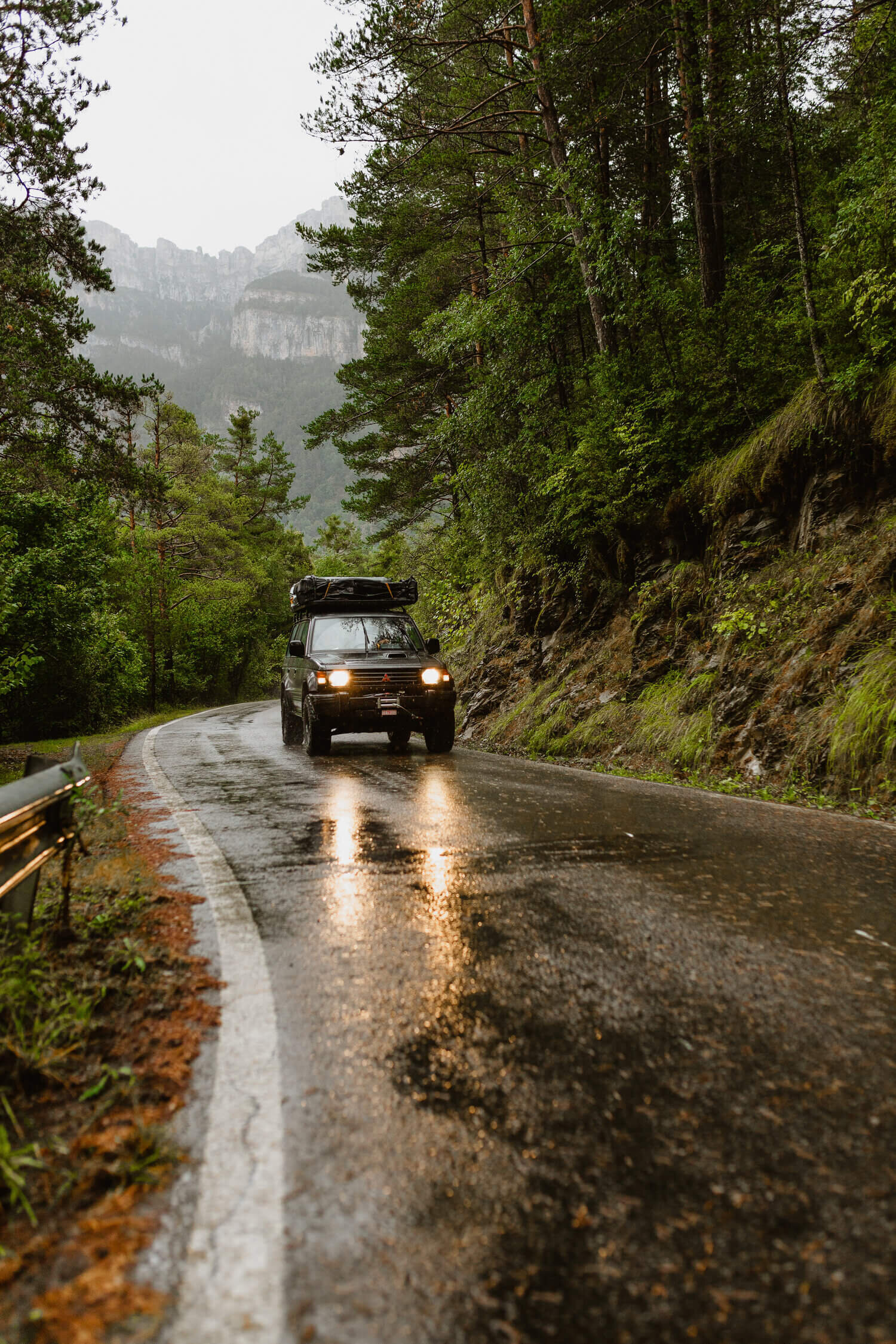 SUV noir sur une route humide en forêt avec des arbres et des montagnes en arrière-plan.