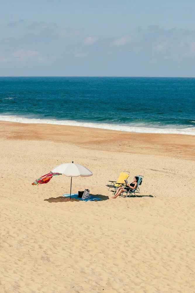 Woman relaxing on a yellow beach chair with a tablet on a sandy beach, under a white umbrella with a colorful towel, near the ocean with waves and a partly cloudy sky.