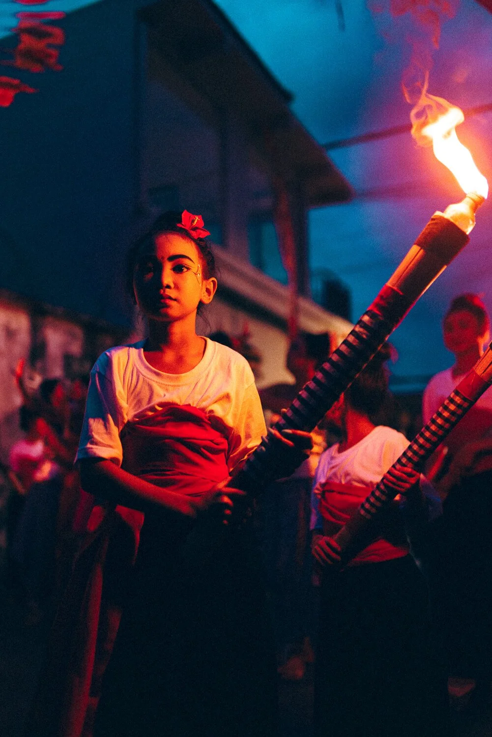 Une jeune fille participe à une parade nocturne, tenant un grand feu d'artifice dans un long tube. La scène est illuminée par la flamme et des lumières colorées, avec plusieurs personnes en arrière-plan.