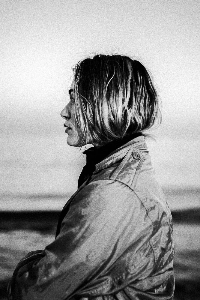 A black-and-white photo of a woman with shoulder-length hair, standing outdoors near water, with her arms crossed, wearing a jacket, in profile view.