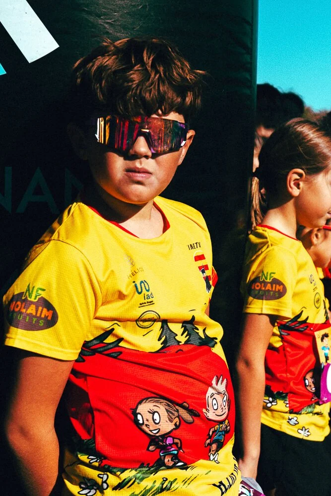 Child with brown hair wearing reflective sunglasses and a yellow and red sports jersey with cartoon characters, standing outdoors.