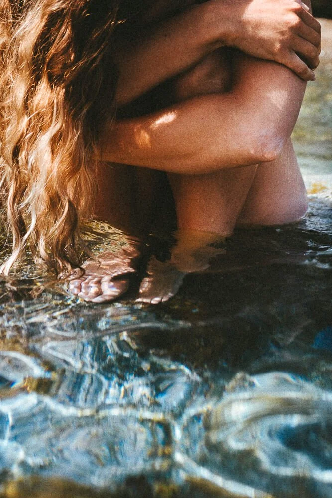 A person with long, curly hair sitting in shallow water, hugging their knees to their chest.