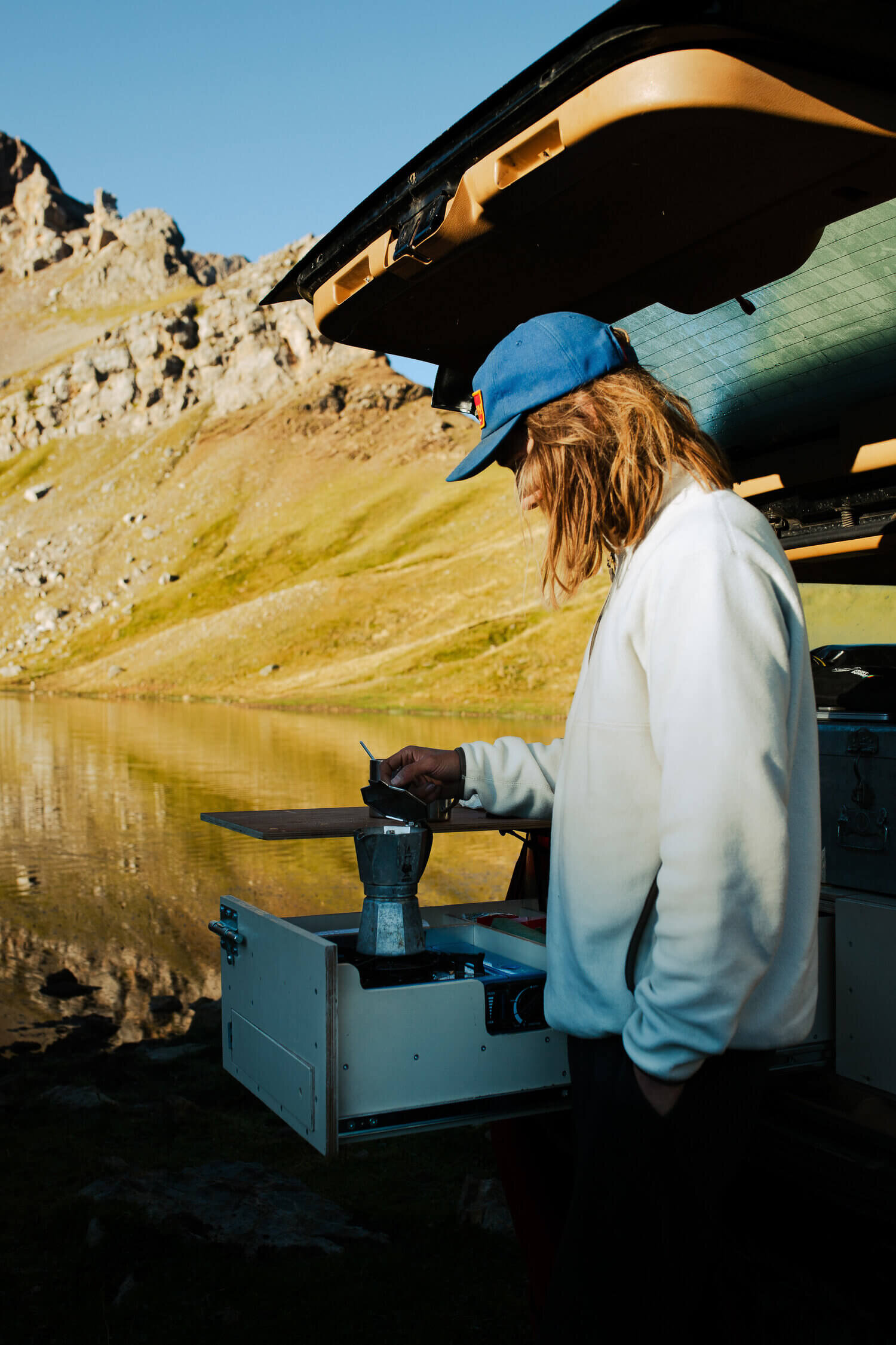 Personne faisant du café avec une cafetière dans le coffre d'une voiture près d'un lac de montagne.