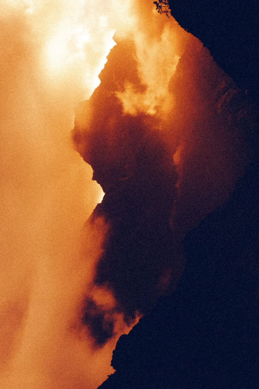 A volcanic eruption with ash and smoke spewing from the volcano's crater under a fiery sky during sunset or sunrise.