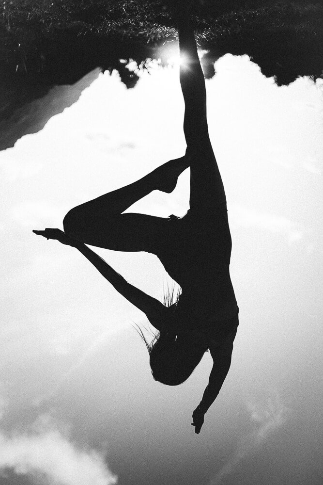 Silhouette of a woman doing a yoga pose outdoors, with a tree and cloudy sky in the background, captured upside down.