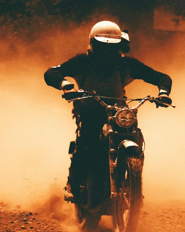 Person riding a motorcycle through dusty terrain, wearing a white helmet, dark clothing, and gloves.