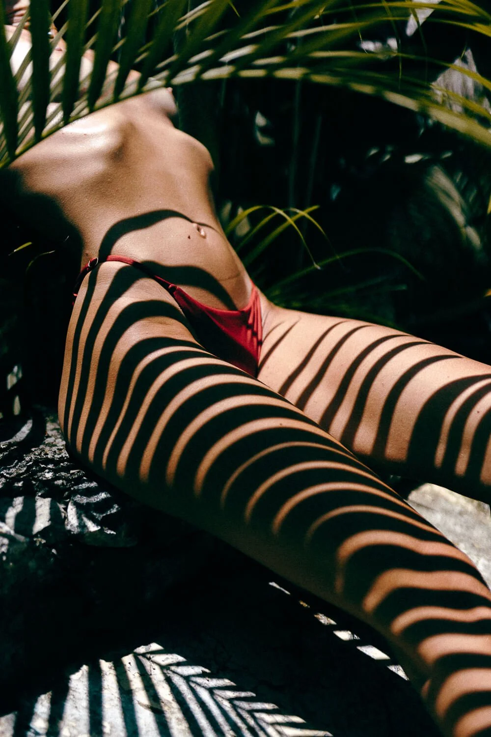 A woman lying on the ground in a tropical setting, partially covered by shadows from nearby palm leaves, wearing red underwear.