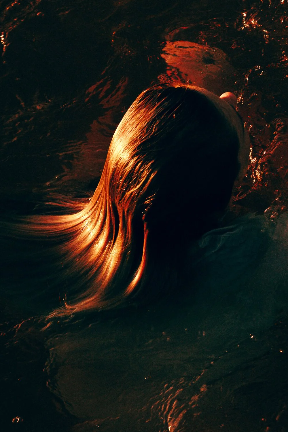 A person with long, flowing hair is submerged in dark water, with sunlight highlighting the hair's texture.
