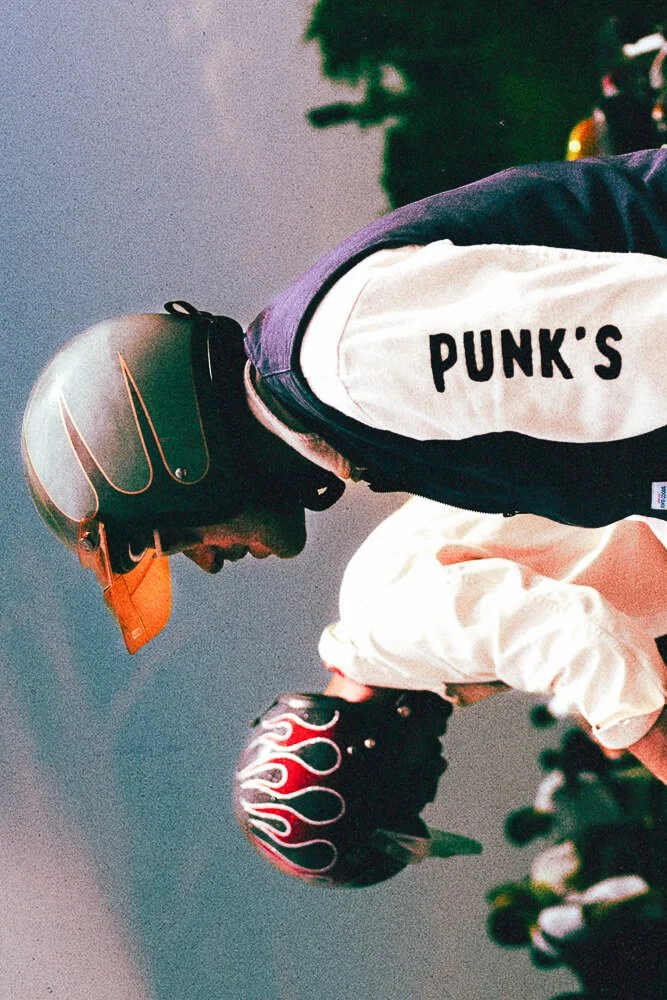 Two individuals wearing helmets, one with a black helmet with flame design and the other with a black helmet with flame graphics, standing outdoors during sunset.