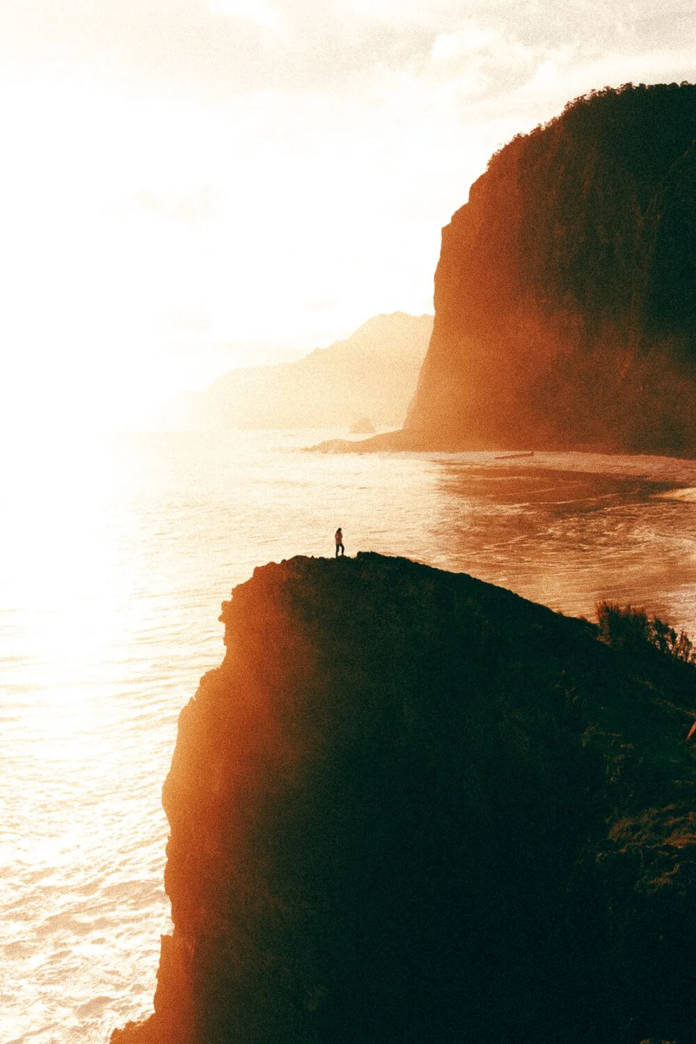 A person standing on a dark cliff overlooking the ocean at sunset with large cliffs in the distance.