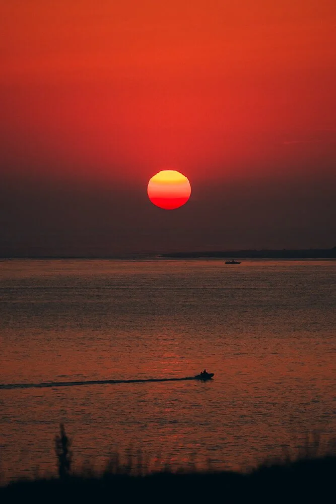 Sunset over a body of water with an orange and pink sky, a boat on the water, and a small vessel creating a wake.