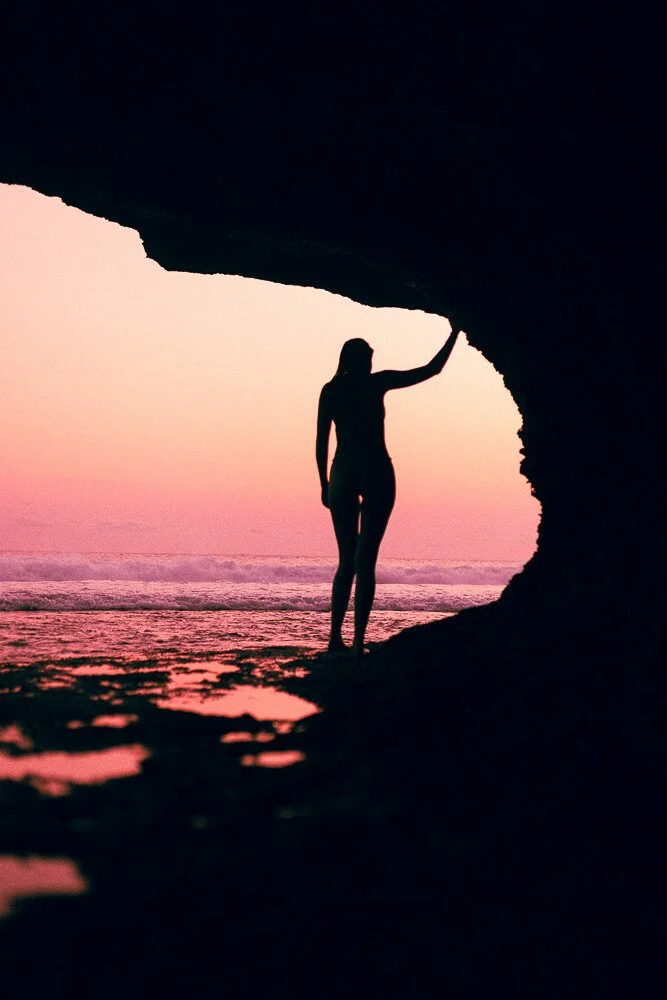 Silhouette of a woman standing inside a cave, holding the cave roof with her hand, with a pink sunset sky and ocean in the background.