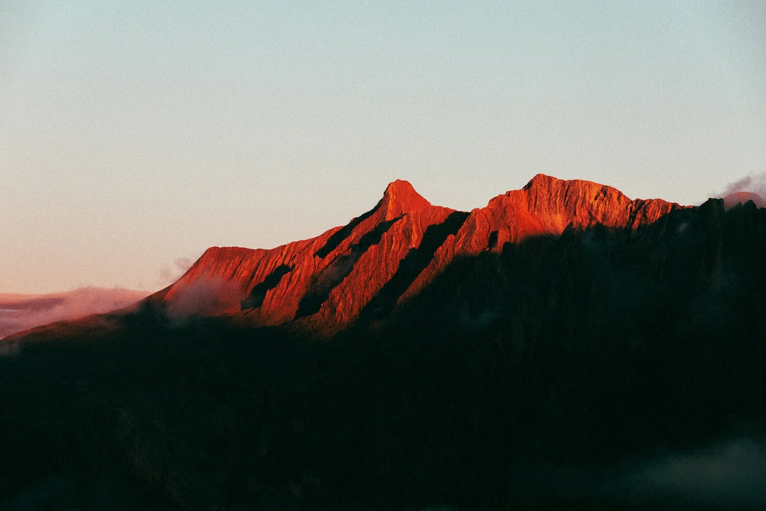 Montagne avec sommets rouges éclairés par le soleil couchant, entourée de brume et de ciel clair.