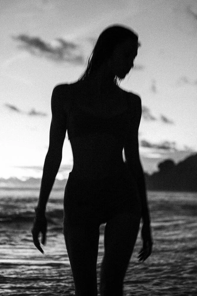 Silhouette of a woman standing in shallow water at the beach during sunset or sunrise with a cloudy sky.