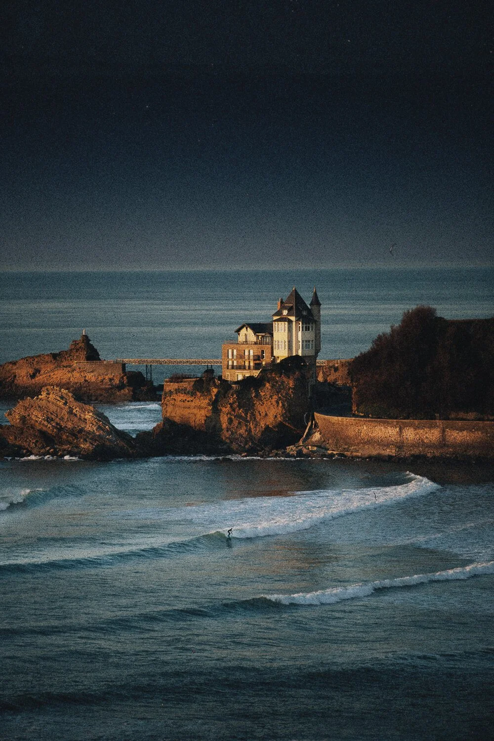 A coastal scene at night featuring a castle-like house on rocky cliffs, with a person surfing in the ocean waves below.