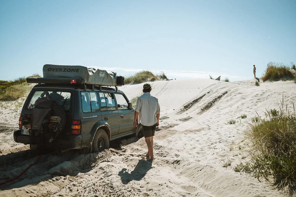 A person walks away from a dark-colored SUV on a sandy beach, with another person and a dog in the distance on the dunes, under a clear blue sky.