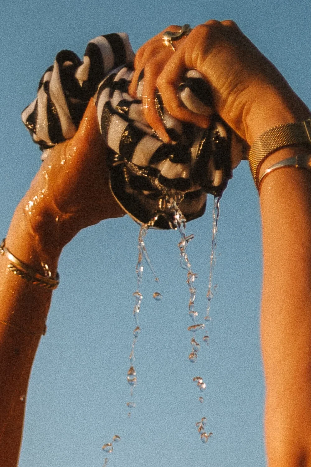 Two hands holding a black and white striped cloth filled with water, with water dripping from the cloth against a clear blue sky background.