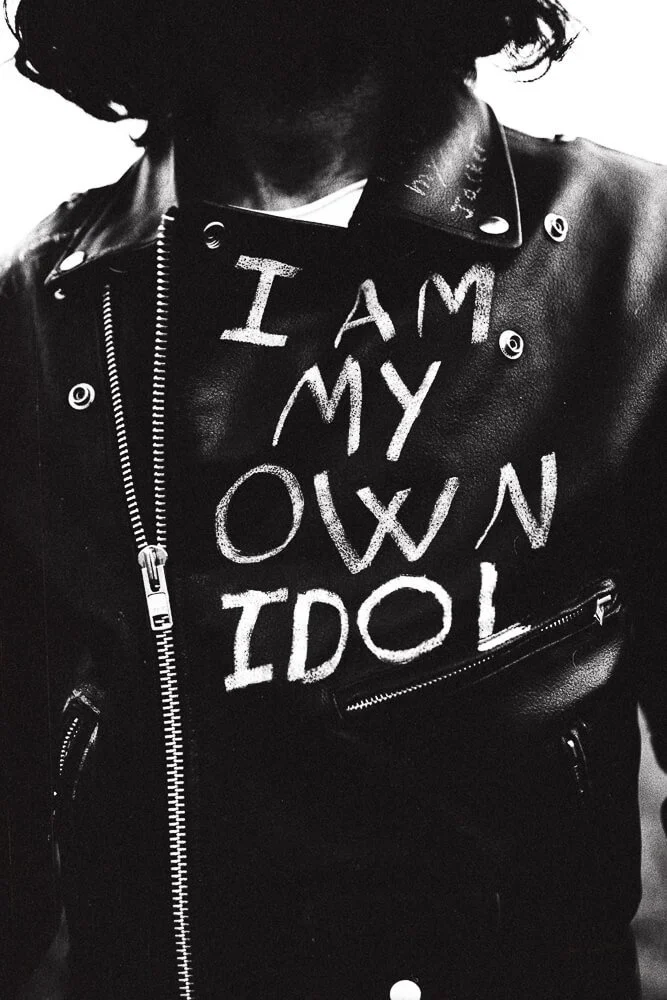 A person wearing a black leather jacket with a distressed white text that reads "I AM MY OWN IDOL."