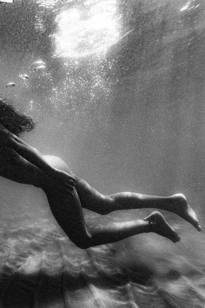 A person swimming underwater, captured in black and white, with their body extended and hair flowing behind them.