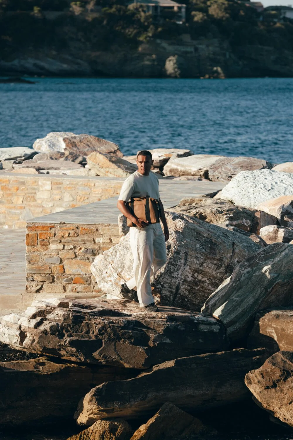 A man standing on large rocks by the water, holding a bag, with a body of water and hills in the background.