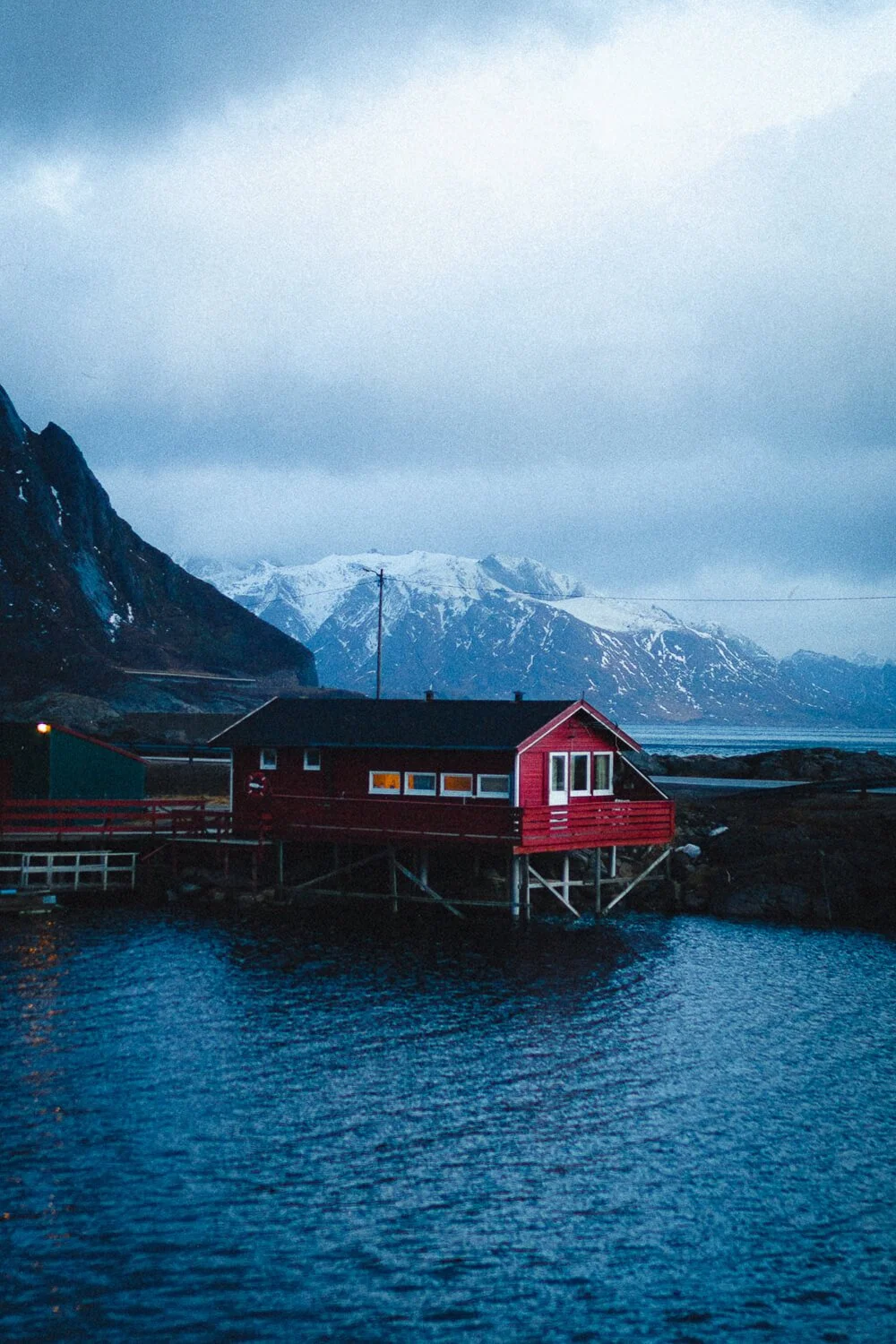 Cabane rouge sur pilotis au bord de l'eau avec montagnes enneigées en arrière-plan, ciel nuageux, ambiance froide et sombre.