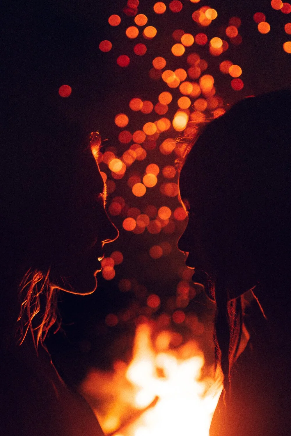 Silhouettes of two women facing each other with a fire behind them, and bokeh-style lights in the background creating a warm, romantic atmosphere.