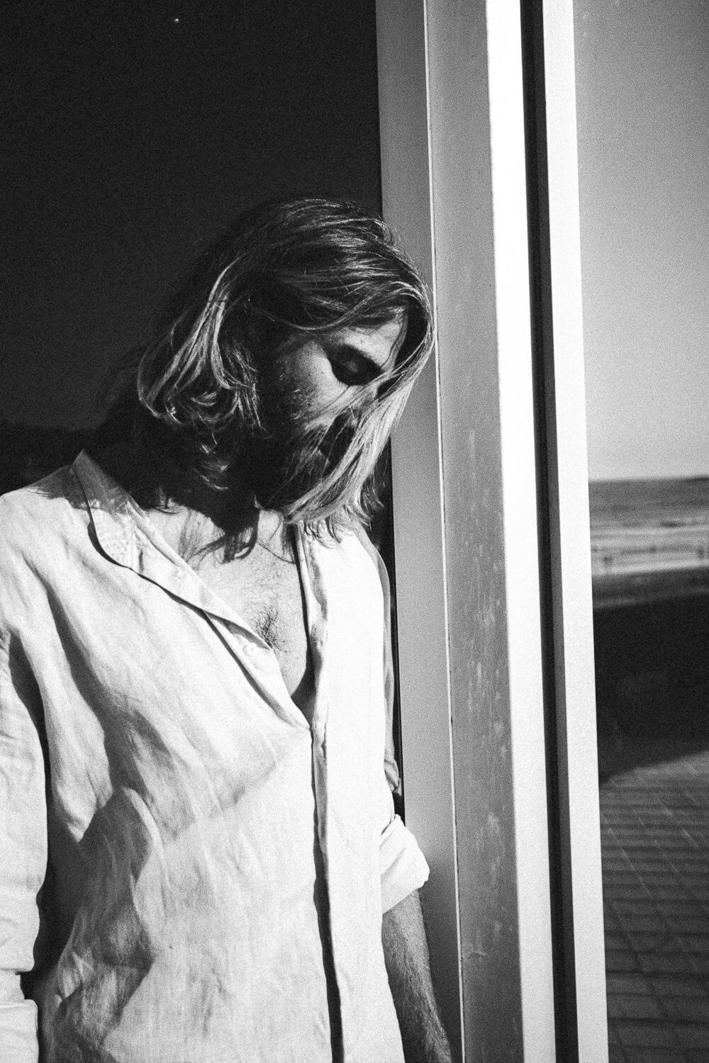 A man with long hair and a beard, wearing a button-up shirt with the top buttons undone, standing by a window looking down, in black and white.