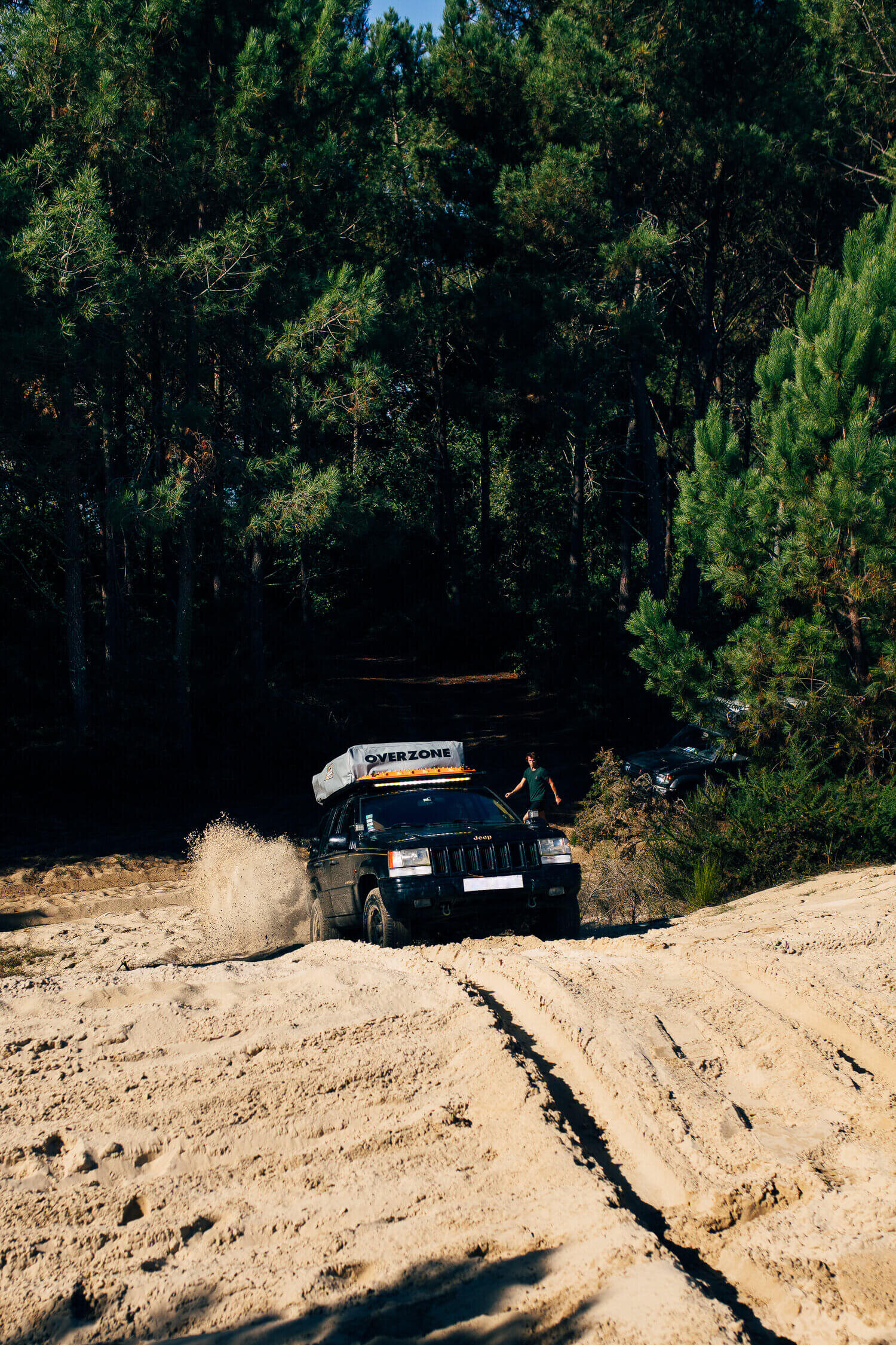 4x4 traversant un terrain sablonneux dans une forêt, avec des arbres verts en arrière-plan. Une personne marche à côté du véhicule. La voiture a un équipement de toit marqué "OVERZONE" et soulève de la poussière.