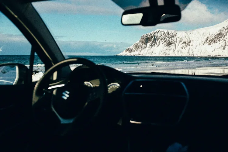 Vue depuis l'intérieur d'une voiture, montrant le tableau de bord, le volant et le miroir, avec une vue sur une plage enneigée et une falaise rocheuse au loin.