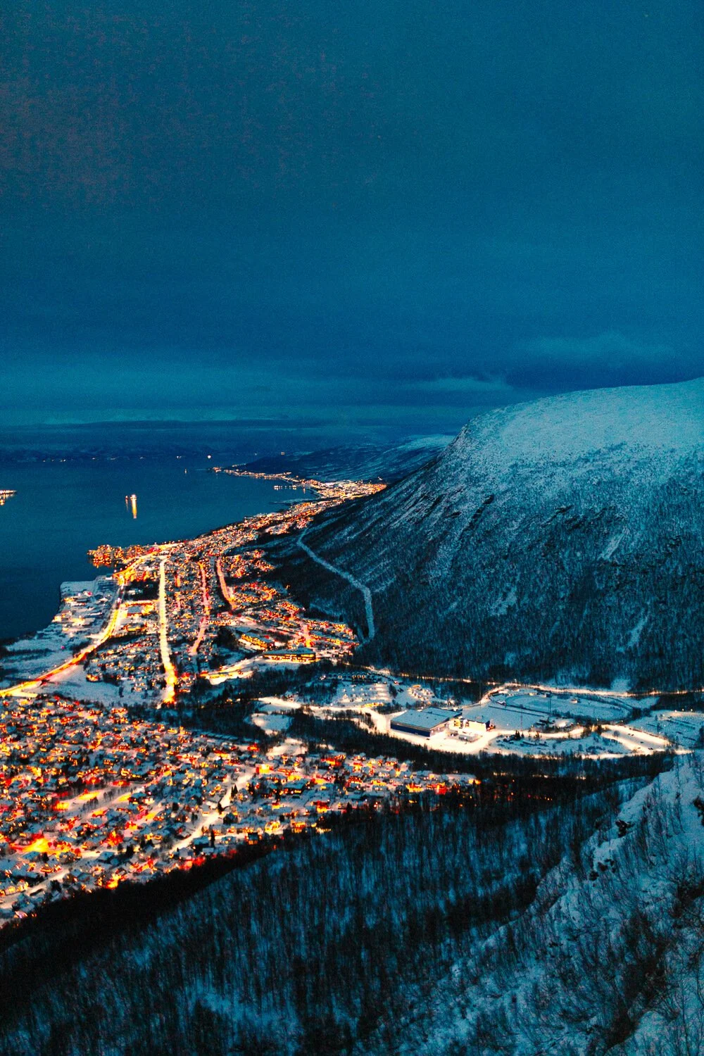 Vue aérienne d'une ville côtière la nuit avec des lumières illuminant les bâtiments et les routes, un lac à proximité, et une montagne enneigée dans le paysage.