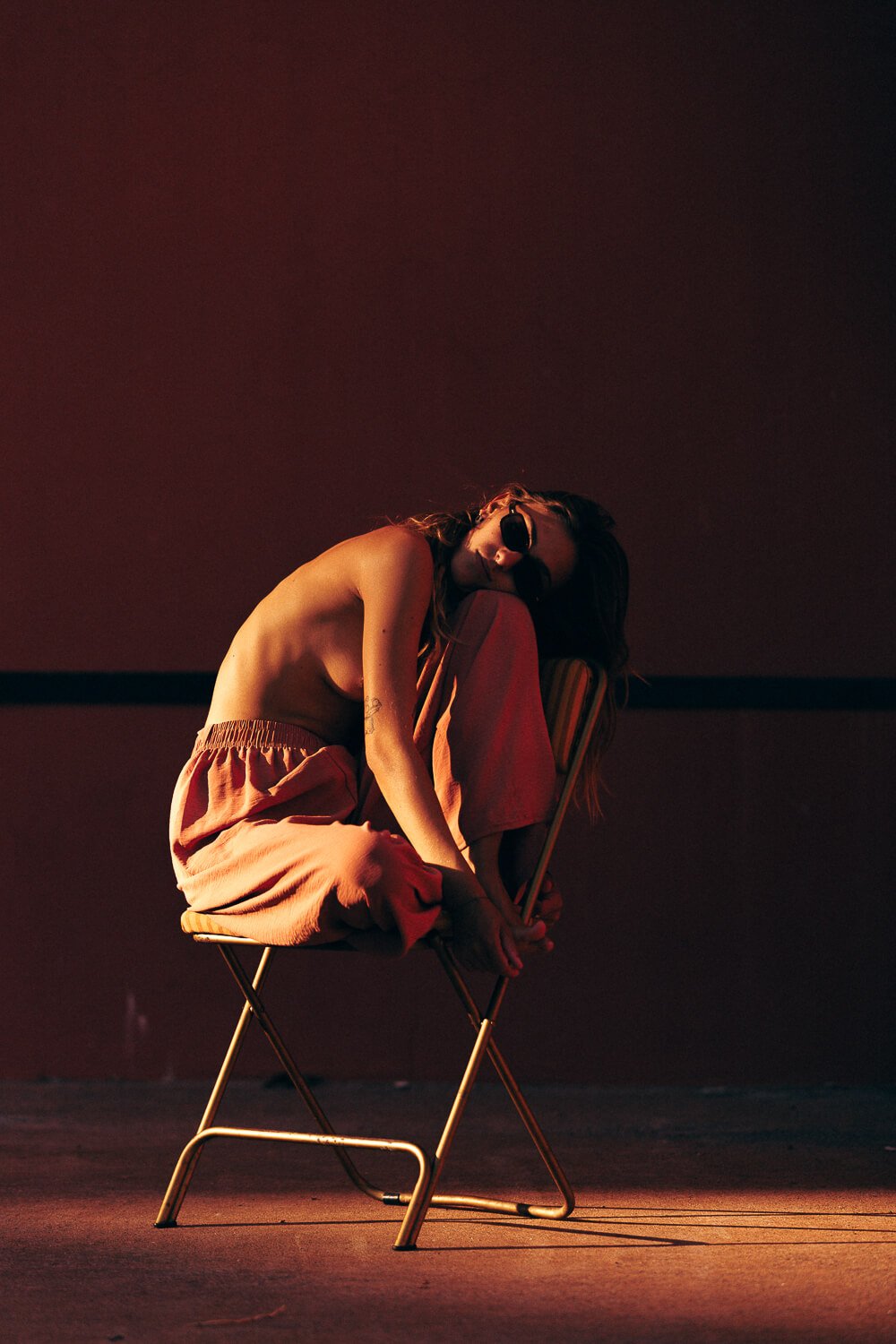 A woman with long hair wearing sunglasses, sitting on a yellow folding chair, topless with her knees drawn up, pink pants, in a dimly lit room with reddish lighting.