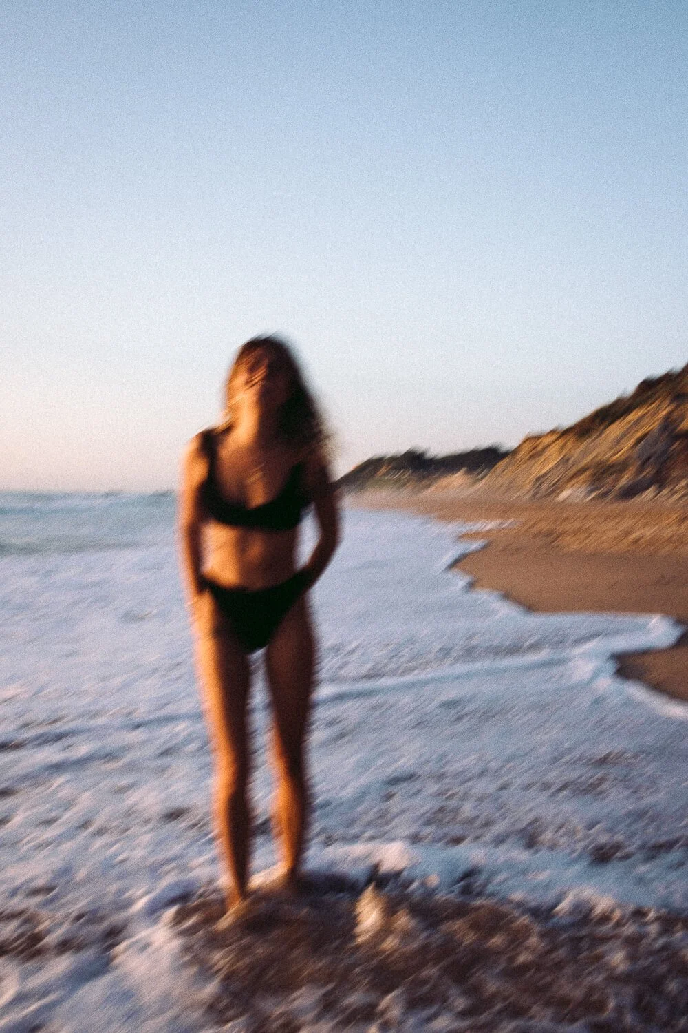 Blurry photo of a woman in a black bikini standing on a sandy beach near the water, with cliffs in the background and the sky above.