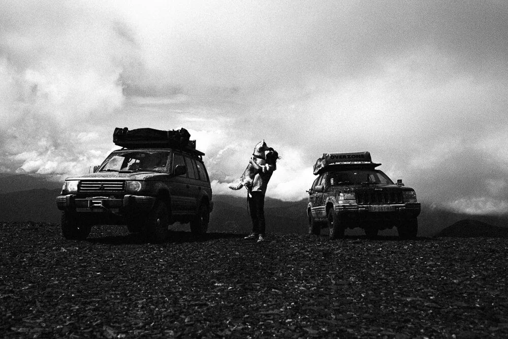 A person holding a dog near two parked SUVs in a rugged outdoor landscape with mountains and cloudy skies in the background.