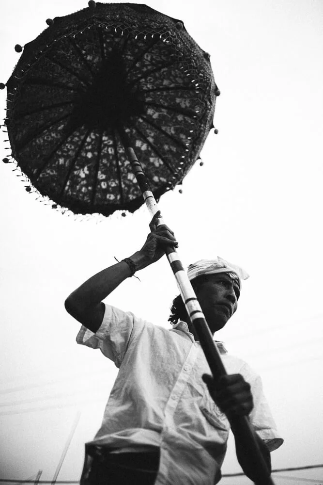 A man holding a decorative umbrella while wearing a white shirt and head wrap, against a plain sky background.
