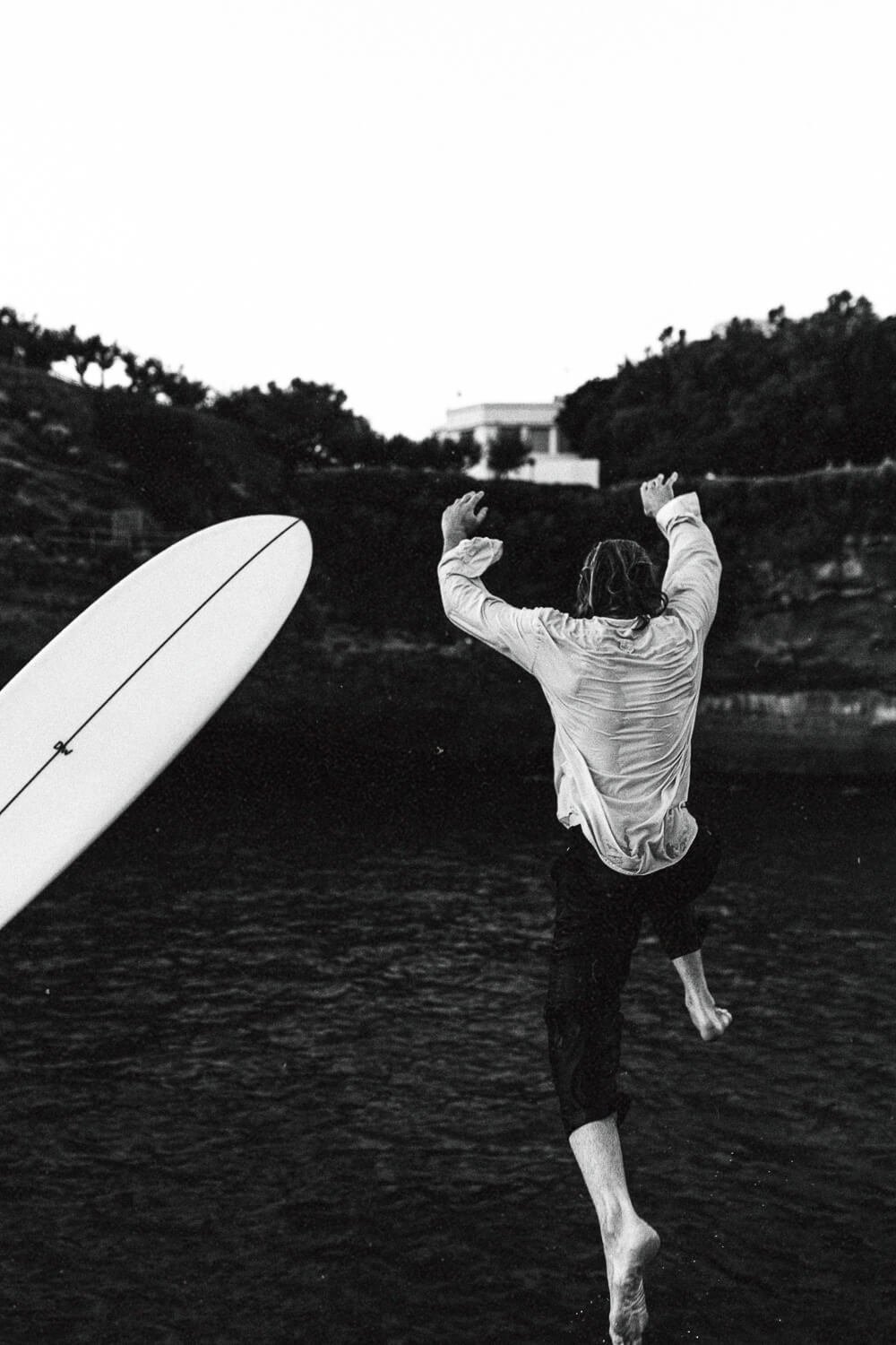 A person jumping into water with a surfboard nearby, black and white photo, trees and building in the background.