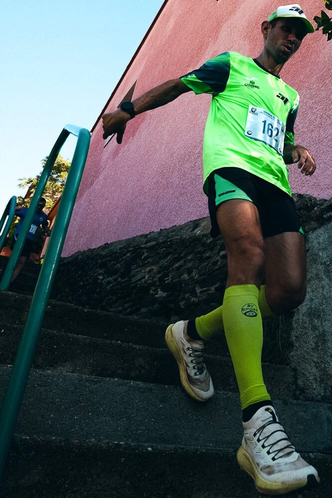 A male runner in a bright green and black athletic outfit, wearing yellow compression socks and white running shoes, descending outdoor stairs during a race.