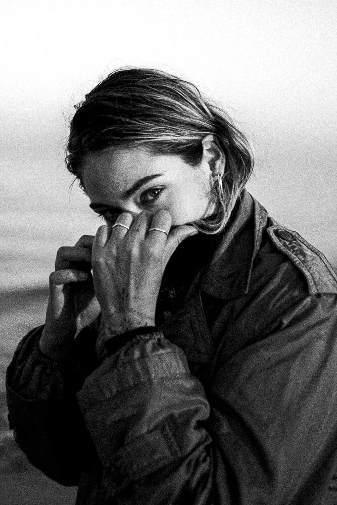 Black and white photo of a woman with short hair, wearing a jacket, covering part of her face with her hands, looking at the camera.