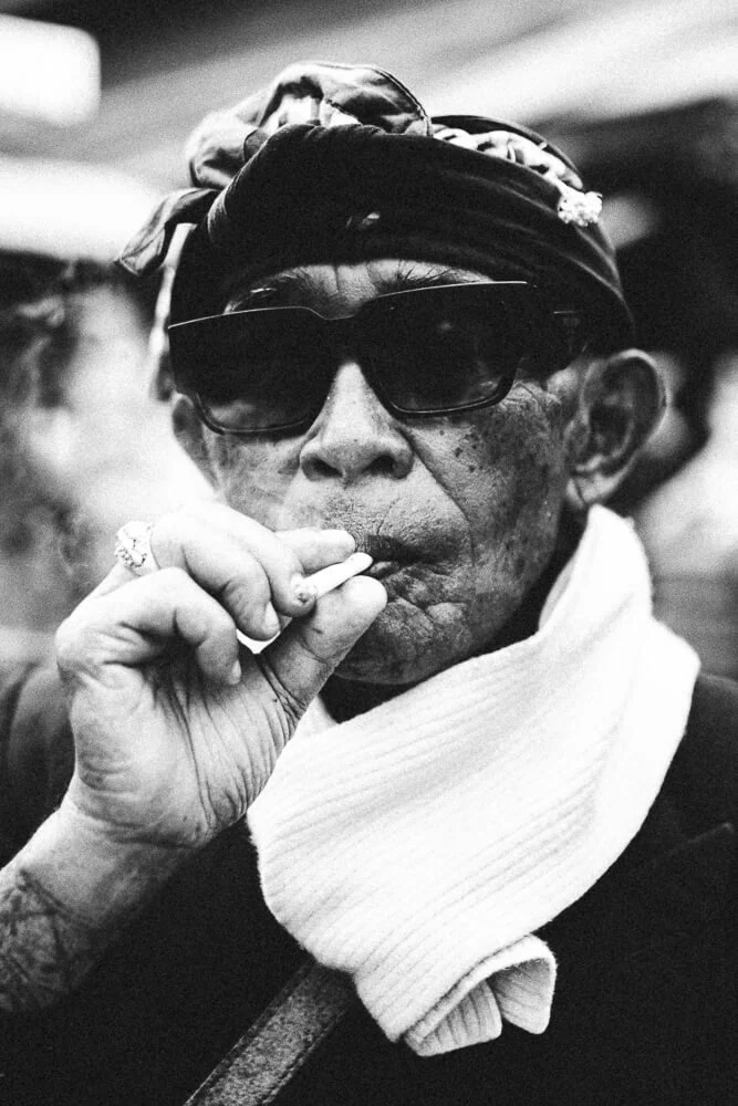 Black and white photo of a person wearing dark sunglasses, a bandana on their head, and a high-collared shirt, smoking a cigarette.