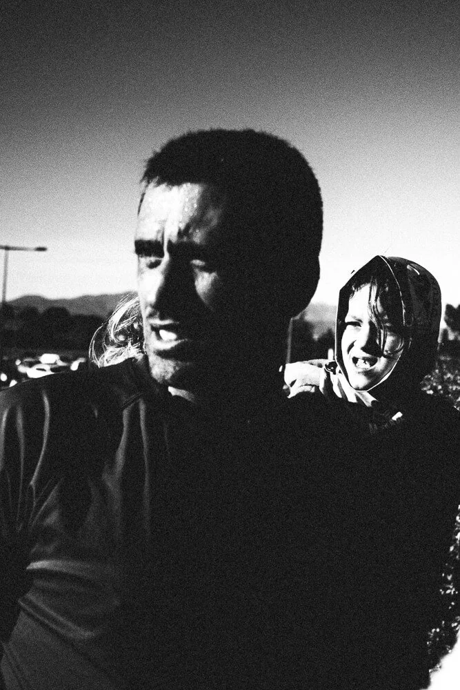 Black and white photo of two people outdoors, one man in the foreground looking to the side with short hair and serious expression, and a child in the background smiling and wearing a hood.