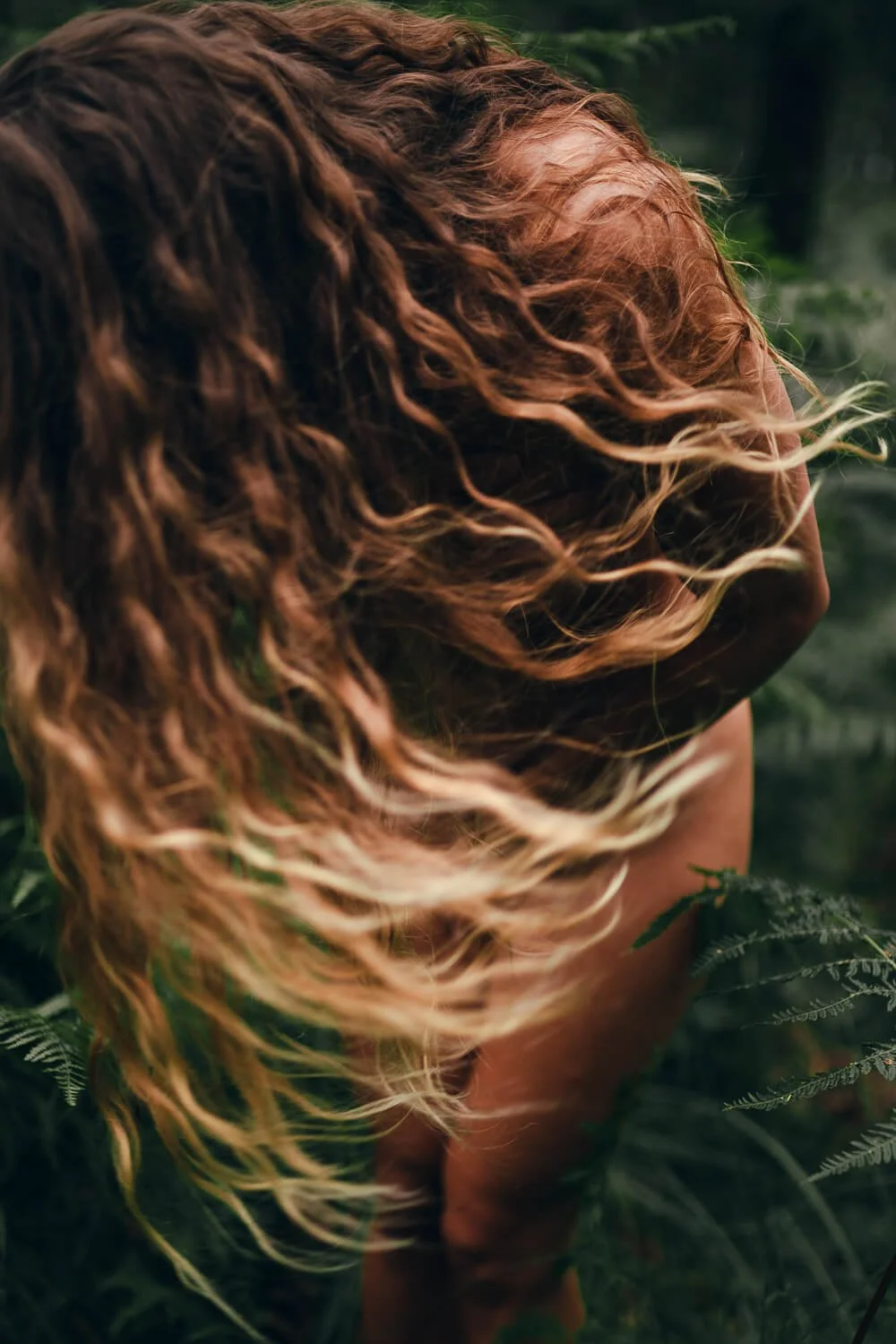 A close-up of a person with curly, reddish-brown hair, standing amidst green foliage outdoors.