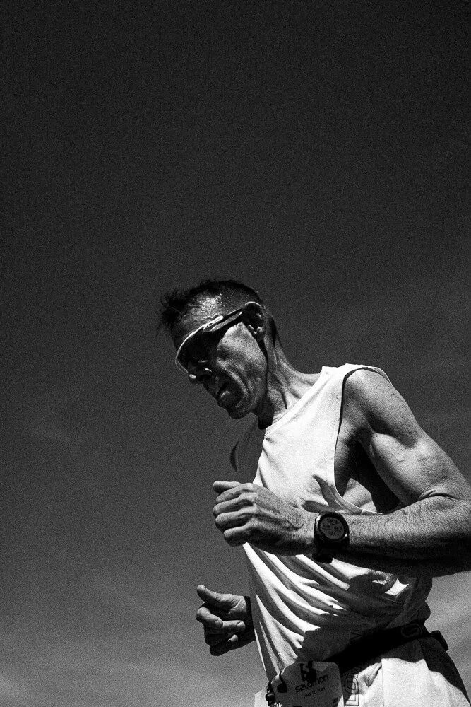 A male runner wearing sunglasses and a white tank top, running outdoors against a dark sky.