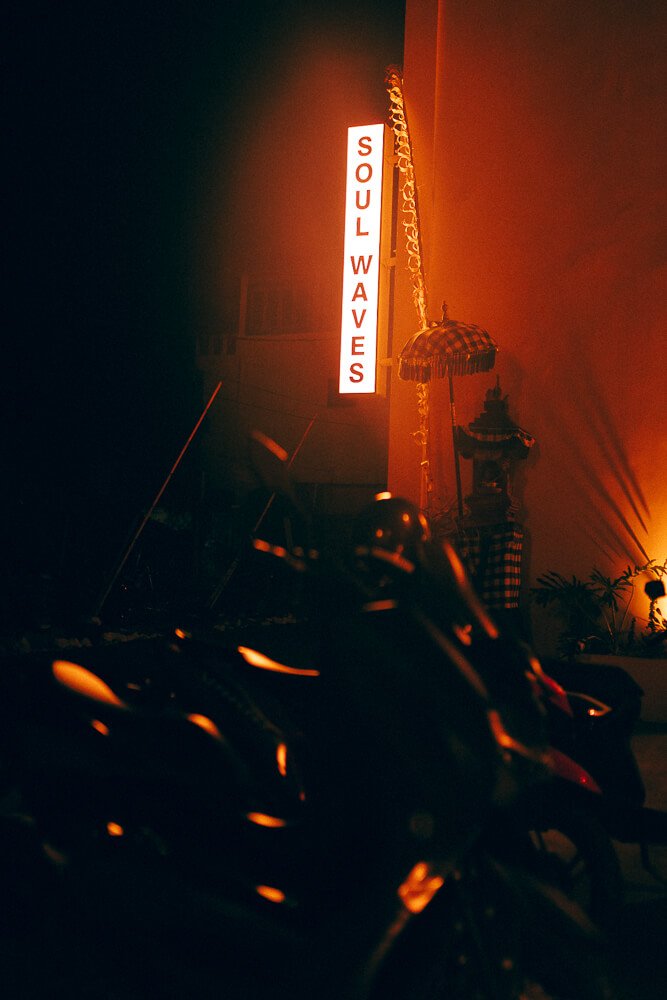 Neon sign reading 'Soul Waves' illuminates a dimly lit room with motorcycles in the foreground and a table with a checked tablecloth and a decorative umbrella in the background.